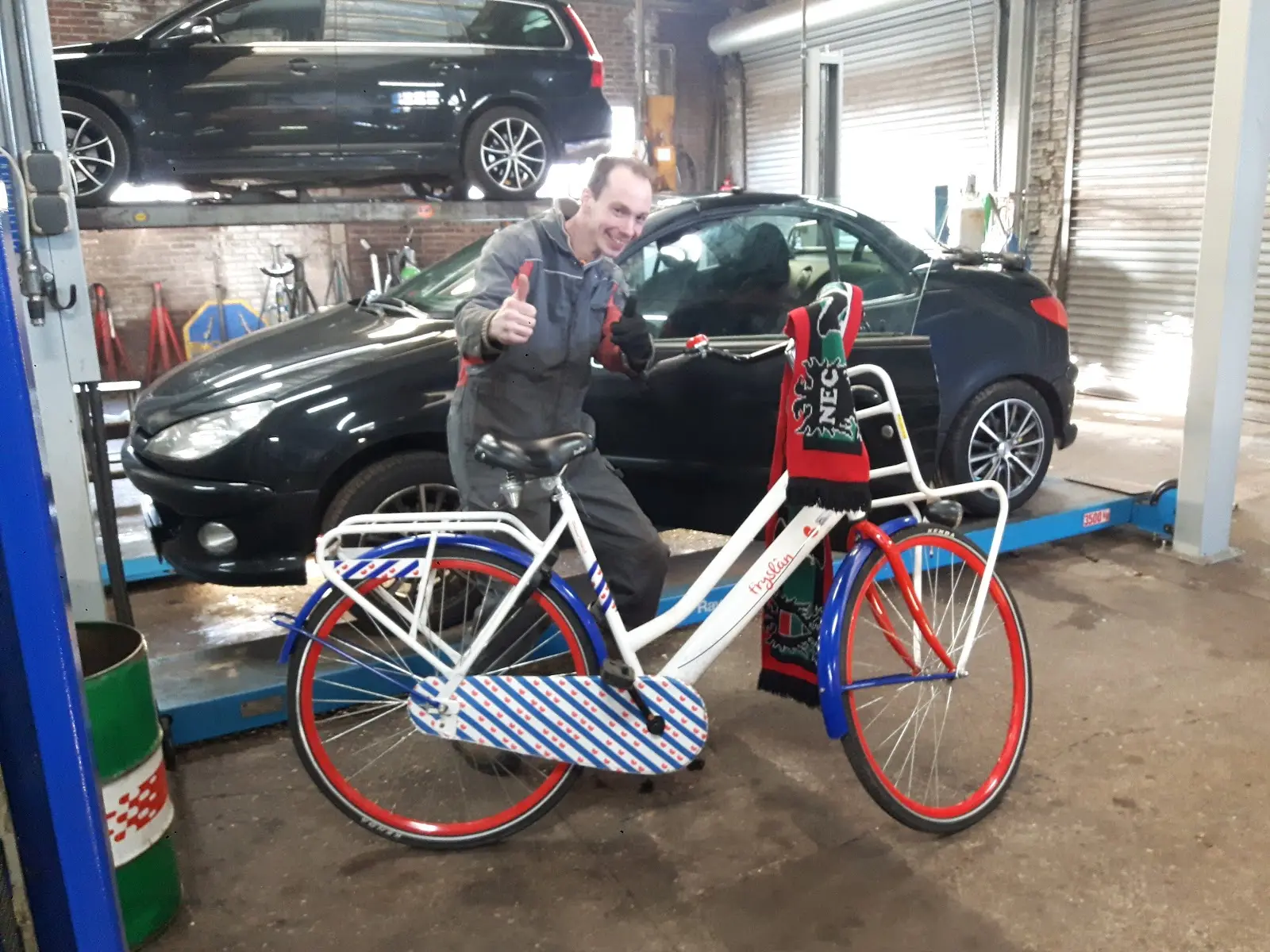 Monteur steekt zijn duim op in de werkplaats, met auto's op de brug en een fiets in Friese kleuren op de voorgrond